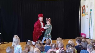 Dzieci zostało zaproszone przez aktora na scenę. Aktor w czerwonym stroju lokaja, dziecko w zielonej sukience. Na widowni siedzą dzieci z przedszkola.