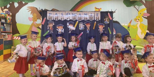 Pasowanie na przedszkolaka dzieci stoją w biretach na tle dekoracji